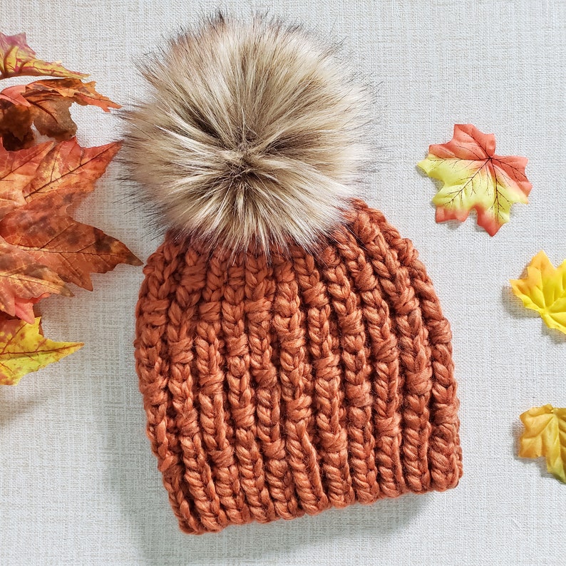 May include: A rust-colored knitted beanie with a fluffy brown pom pom. The beanie is made with a chunky yarn and has a ribbed texture.