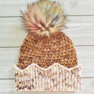 May include: A brown and cream crocheted beanie with a fluffy multi-colored pom pom on top. The beanie has a scalloped brim.