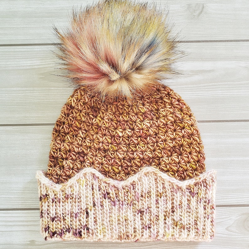 May include: A brown and cream crocheted beanie with a fluffy multi-colored pom pom on top. The beanie has a scalloped brim.