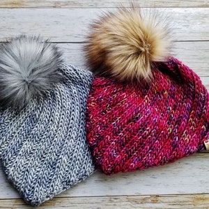 May include: Two knitted winter hats with faux fur pom poms. One hat is gray with a gray pom pom, and the other is pink with a brown pom pom.