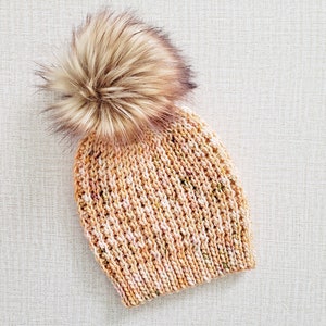 May include: A knitted beanie with a fluffy pom pom in shades of peach, pink, and brown.