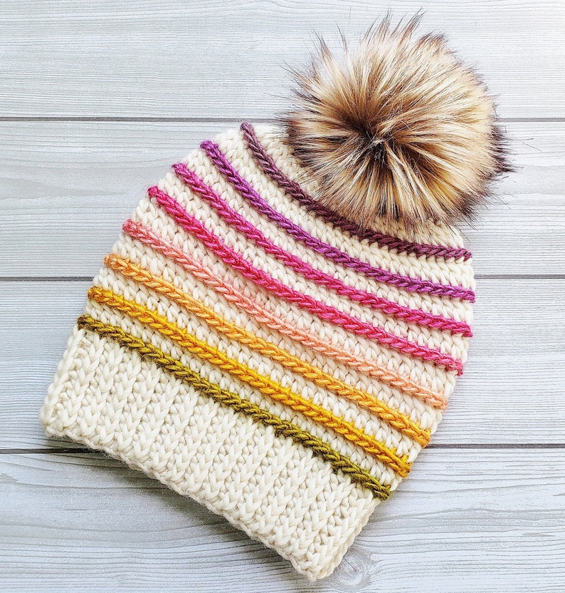 Pode incluir: Um gorro branco de croch&ecirc; com um padr&atilde;o listrado colorido em tons de rosa, laranja, amarelo e verde. O gorro tem um pompom marrom felpudo no topo.
