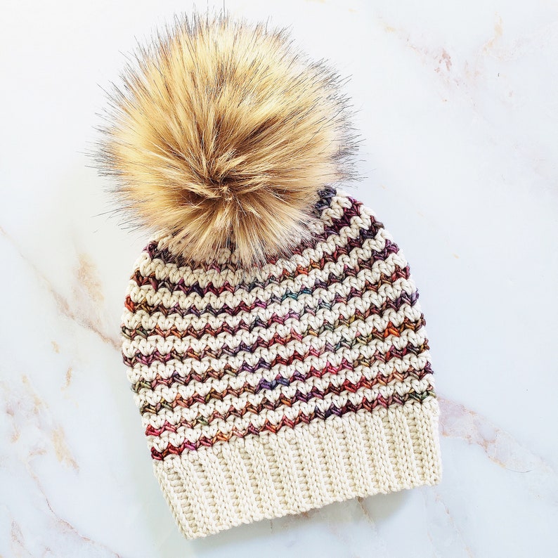 May include: A crocheted beanie with a multi-colored chevron pattern and a fluffy brown pom pom.