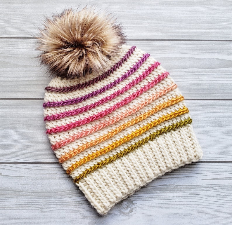 Pode incluir: Um gorro branco de croch&ecirc; com um padr&atilde;o listrado em tons de rosa, laranja, amarelo e verde. O gorro tem um pompom marrom felpudo no topo.