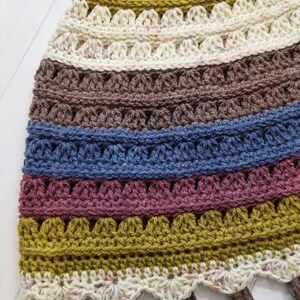 May include: A crocheted scarf with a multi-colored striped pattern in shades of brown, blue, pink, and green. The scarf has a scalloped edge and brown fringe.