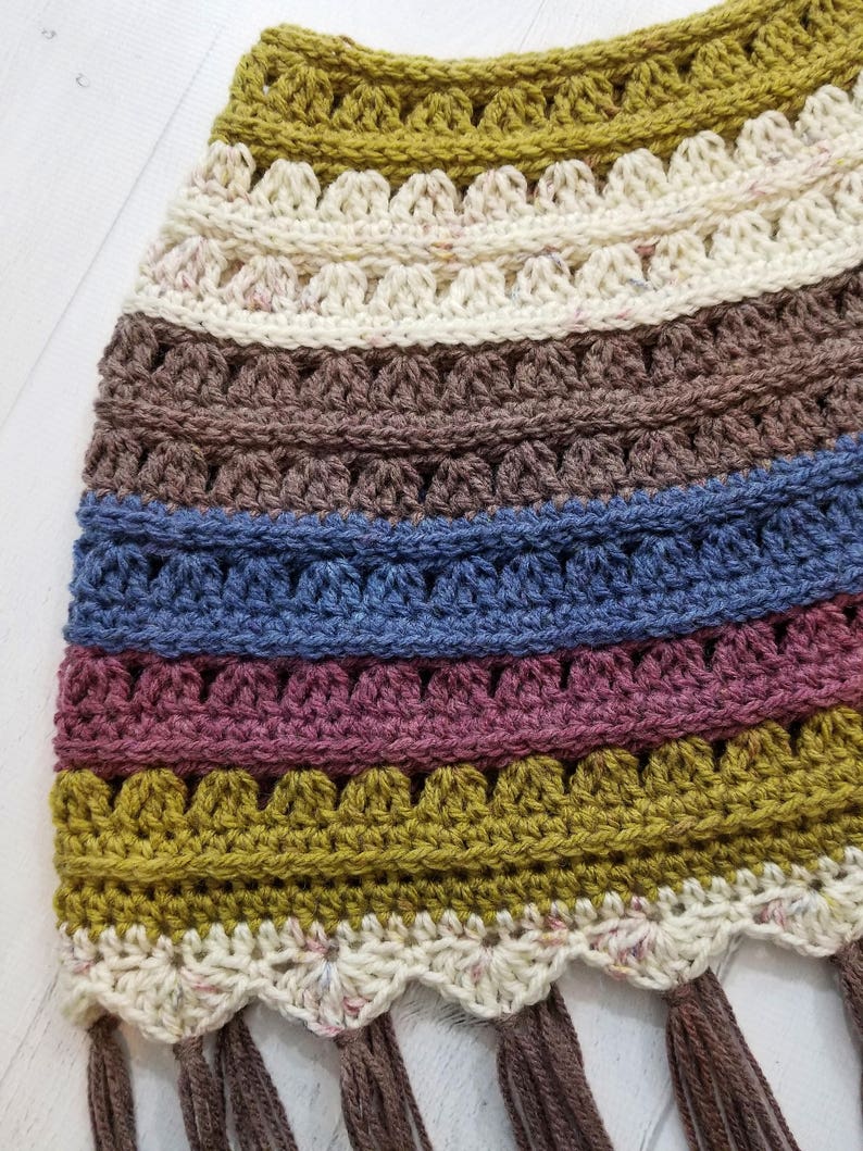 May include: A crocheted scarf with a multi-colored striped pattern in shades of brown, blue, pink, and green. The scarf has a scalloped edge and brown fringe.