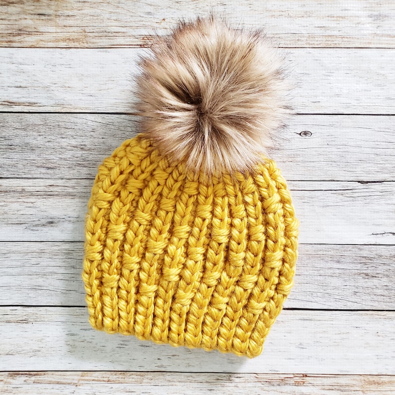May include: A yellow knitted beanie with a brown faux fur pom pom.