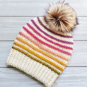 Pode incluir: Um gorro branco de croch&ecirc; com um padr&atilde;o listrado colorido e um pompom de pele sint&eacute;tica marrom.