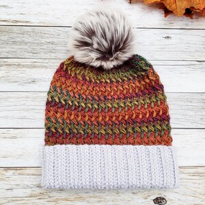 May include: A handmade knit hat with a light gray ribbed brim and a colorful woven pattern in shades of red, orange, green, and yellow. The hat is topped with a fluffy, faux fur pom-pom in brown and white.