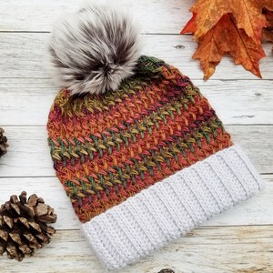 May include: A hand-knitted winter hat with a gray ribbed brim and a colorful woven pattern in shades of orange, red, green, and yellow. The hat is topped with a large, fluffy, gray and brown faux fur pom-pom. The hat is displayed on a light wood surface.