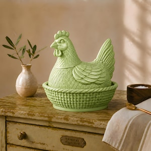 May include: A light green ceramic hen-shaped egg holder with a basket-weave base. The hen is perched on a distressed wooden surface, next to a small vase with greenery. A small wooden bowl with salt and a gold spoon are also present.