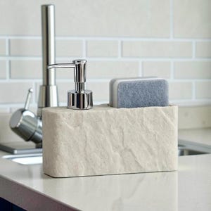 May include: A cream-colored soap dispenser and sponge holder set. The dispenser has a chrome pump, and a gray and white sponge sits in the holder. The set is made of a stone-like material and sits on a countertop.
