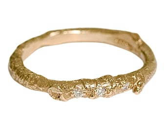 May include: A gold-colored ring with a textured, twig-like design. The ring features three small, sparkling gemstones set along the band. The ring is set against a white background, highlighting its intricate details.