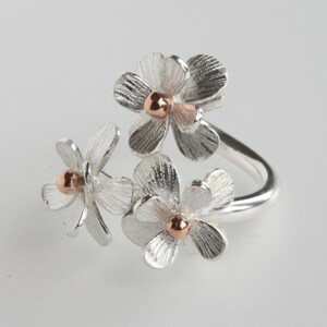 May include: A silver ring with three flower motifs. Each flower has textured petals and a small, copper-coloured centre. The ring band curves around, creating a delicate, nature-inspired design.