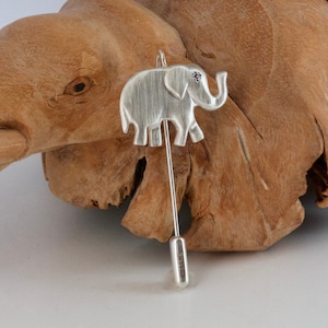 May include: A silver elephant lapel pin with a small red gem eye. The pin is made of silver and has a long, thin needle. The elephant is detailed with etched lines. The pin is displayed on a wooden surface.