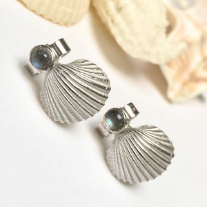 May include: A pair of silver seashell stud earrings with a small, round, iridescent stone set above each shell. The earrings are displayed on a white surface with a blurred seashell in the background.