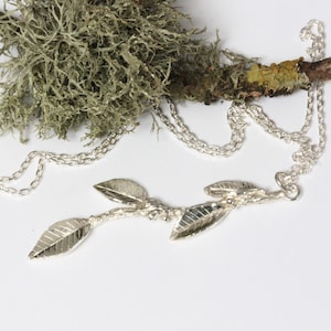May include: A silver-colored necklace featuring a delicate leaf design. The pendant has three detailed leaves attached to a branch-like structure. The chain is a simple link style. The background includes a branch with green and brown moss.