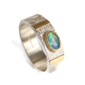May include: A silver ring with gold detailing and an oval opal gemstone. The ring features a textured band and a gold bezel setting. The opal displays a mix of blue, green, and yellow colours.