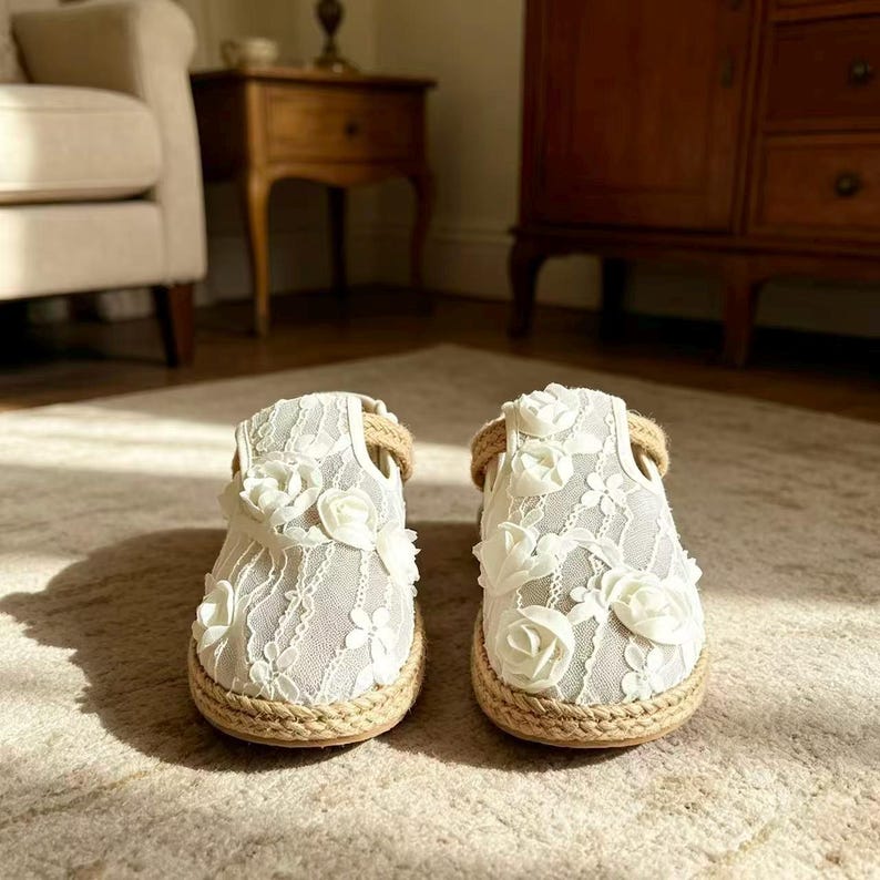 K&ouml;nnte beinhalten: Ein Paar wei&szlig;e Baby-Schuhe aus Spitze mit floralen Akzenten. Die Schuhe haben eine Sohle aus geflochtenem Seil und einen Riemen um den Kn&ouml;chel. Die Schuhe sind etwa 10 cm lang.