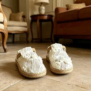 K&ouml;nnte beinhalten: Paar wei&szlig;e Espadrilles-Schuhe mit floraler Spitzenauflage und Juteseilbesatz. Die Schuhe haben eine runde Spitze und eine flache Sohle. Der Hintergrund ist neutral.