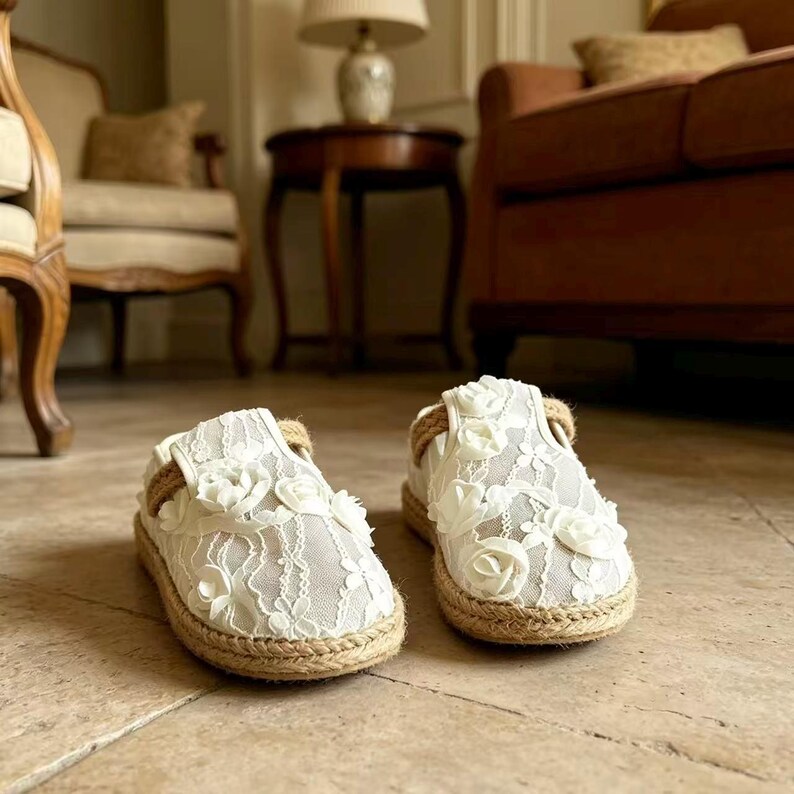K&ouml;nnte beinhalten: Paar wei&szlig;e Espadrilles-Schuhe mit floraler Spitzenauflage und Juteseilbesatz. Die Schuhe haben eine runde Spitze und eine flache Sohle. Der Hintergrund ist neutral.
