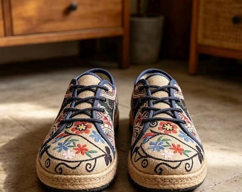 Könnte beinhalten: Ein Paar flache Sneaker mit einem Obermaterial aus beigem Webstoff und aufwendigen Blumenstickereien in Rot, Blau und Grün. Die Schuhe haben dunkelblaue Schnürsenkel, eine abgerundete Zehenpartie und eine mit Jute umwickelte Sohlenkante.