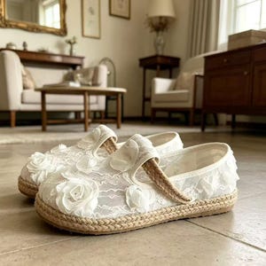 K&ouml;nnte beinhalten: Ein Paar wei&szlig;e Mary Jane-Schuhe aus Spitze mit floralen Verzierungen. Die Schuhe haben eine gewebte Jute-Sohle und einen gewebten Riemen. Der Hintergrund zeigt eine Wohnzimmereinrichtung mit M&ouml;beln.