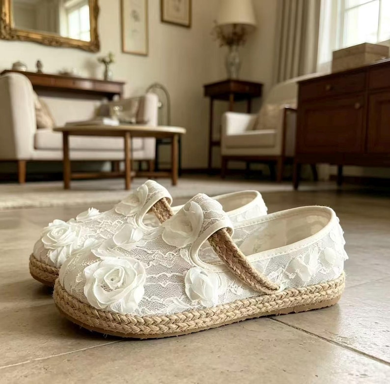 K&ouml;nnte beinhalten: Ein Paar wei&szlig;e Mary Jane-Schuhe aus Spitze mit floralen Verzierungen. Die Schuhe haben eine gewebte Jute-Sohle und einen gewebten Riemen. Der Hintergrund zeigt eine Wohnzimmereinrichtung mit M&ouml;beln.