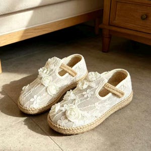 K&ouml;nnte beinhalten: Ein Paar wei&szlig;e Mary Jane-Schuhe aus Spitze mit floralen Verzierungen. Die Schuhe haben eine gewebte Jute-Sohle und einen Riemen. Die Schuhe sind f&uuml;r ein Kind konzipiert.