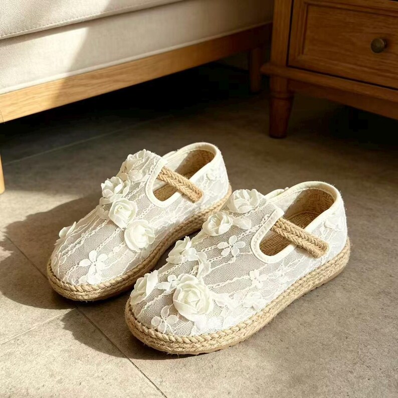 K&ouml;nnte beinhalten: Ein Paar wei&szlig;e Mary Jane-Schuhe aus Spitze mit floralen Verzierungen. Die Schuhe haben eine gewebte Jute-Sohle und einen Riemen. Die Schuhe sind f&uuml;r ein Kind konzipiert.
