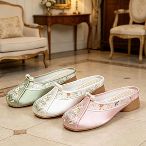 May include: Three slip-on shoes in light green, white, and pink satin. Each shoe features a square toe, a low block heel, a single white pearl accent at the centre front, and delicate floral embroidery on the upper and side panels.