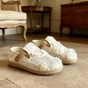 K&ouml;nnte beinhalten: Ein Paar wei&szlig;e Spitzenschuhe mit floralen Akzenten. Die Schuhe haben eine geflochtene Jutesohle und einen geflochtenen Riemen. Diese Kinderschuhe haben ein zartes, feminines Design.