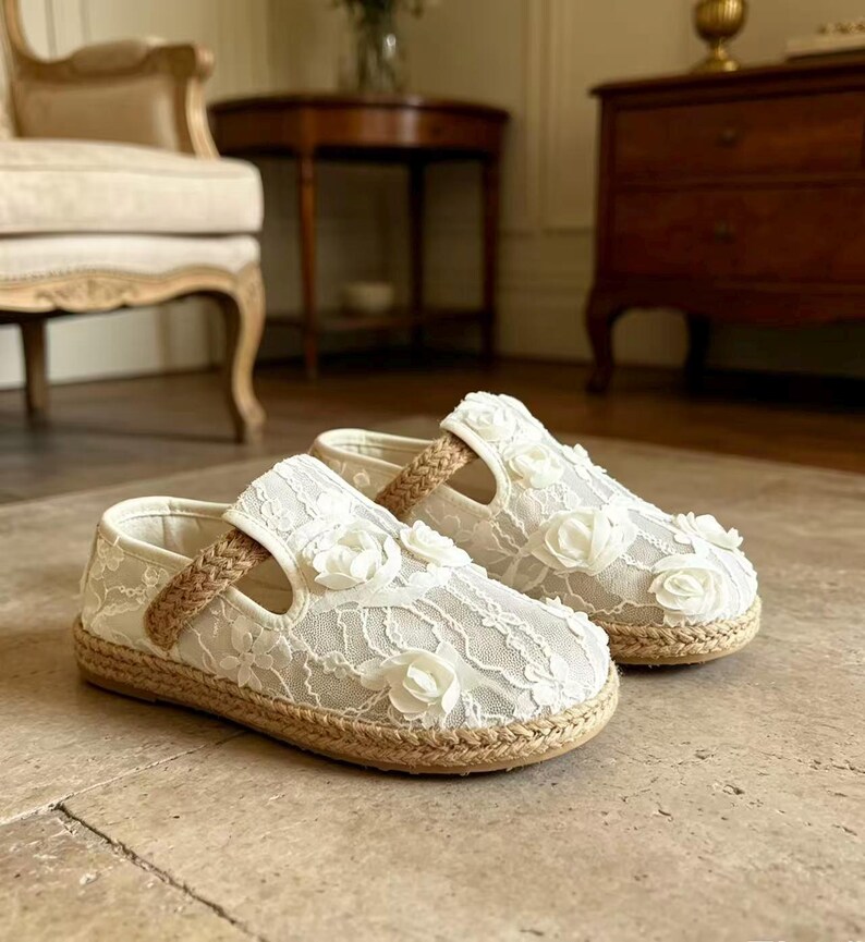 K&ouml;nnte beinhalten: Ein Paar wei&szlig;e Spitzenschuhe mit floralen Akzenten. Die Schuhe haben eine geflochtene Jutesohle und einen geflochtenen Riemen. Diese Kinderschuhe haben ein zartes, feminines Design.