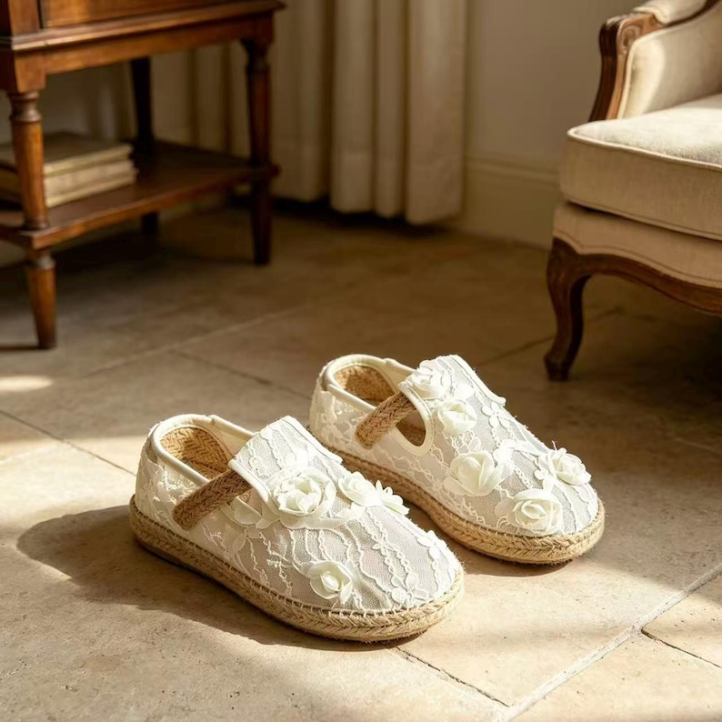 K&ouml;nnte beinhalten: Ein Paar wei&szlig;e Espadrilles-Flats mit floraler Spitze und dekorativen wei&szlig;en Stoffrosen. Die Schuhe haben eine gewebte Jutesohle und einen beigefarbenen Riemen. Die Schuhe werden auf einem gefliesten Boden pr&auml;sentiert.