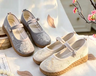 Könnte beinhalten: Zwei Paar Slipper mit Mary-Jane-Riemen und Knotenverschluss. Ein Paar ist hellgrau, das andere beige. Beide haben eine weiße Blumenstickerei an der Spitze und an der Seite sowie eine Zwischensohle in Jute-Optik und eine flache Sohle.