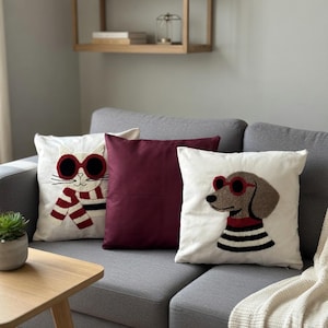 Op de afbeelding: Een grijze bank met drie sierkussens. Twee kussens hebben geborduurde dierenontwerpen: een kat met een zonnebril en een sjaal, en een hond met een zonnebril en een gestreept shirt. Er is ook een effen bordeauxrood kussen.