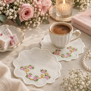 May include: Two round, scalloped-edge white fabric coasters featuring an embroidered design of small pink flowers and yellow vines. One coaster sits beneath a white fluted ceramic cup filled with dark coffee, while the other lies empty.