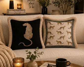 May include: Two square throw pillows with fringe trim. One pillow is black with a single embroidered leopard graphic. The other pillow is cream-colored with a repeating pattern of small leopards and a dark border with a white dot design.
