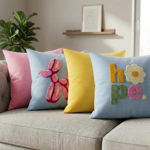 Puede incluir: Un conjunto de cojines decorativos en tonos pastel. Incluye un cojín rosa, un cojín azul claro con un perro globo, un cojín amarillo y un cojín azul claro con la palabra "hope" y una flor.
