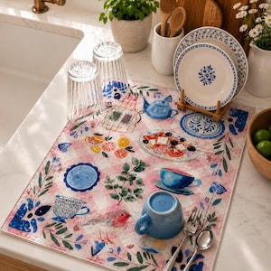 Könnte beinhalten: Eine rechteckige Abtropfmatte für die Küche mit rosa-weißem Vichy-Muster, verziert mit Illustrationen eines Rotkehlchens, Zitrusfrüchten, einem Salatteller, Teetassen und blauen Blumenmotiven. Zwei klare gerippte Gläser und eine blaue Tasse stehen darauf.