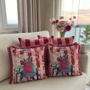 Op de afbeelding: Decoratieve kussens met een rood-roze streepmotief en een olifantenontwerp. De kussens hebben een gefranjerde rand en zijn op een crèmekleurige bank geplaatst. De kussens zijn een decoratief woonaccessoire.