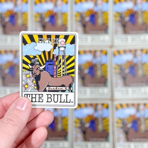 May include: A rectangular sticker featuring a brown bull with a city skyline, water tower, and the text "THE BULL." The design includes yellow sun rays, blue clouds, and white daisies. The sticker is held in a hand.