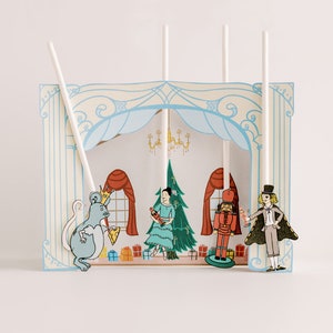 Peut inclure: Un décor de scène en papier avec un fond bleu et blanc. Il y a quatre personnages sur des bâtons : une souris, une ballerine, un casse-noisette et un homme en haut-de-forme. La scène est décorée d’un sapin de Noël et de cadeaux.