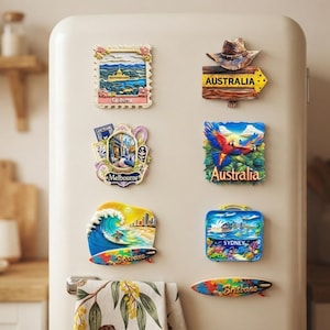 May include: A collection of eight colorful souvenir magnets featuring Australian landmarks and themes, including Canberra, Melbourne, Brisbane, and Sydney, displayed on a cream-colored refrigerator door with a patterned towel hanging below.