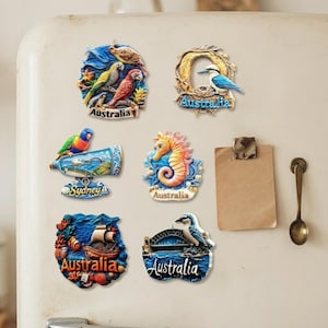 Könnte beinhalten: Sechs dekorative Kühlschrankmagnete mit australischen Motiven, darunter Papageien, ein Kookaburra, ein Seepferdchen, ein Schiff und das Sydney Opera House, mit dem Schriftzug Australia oder Sydney auf jedem Teil, angeordnet auf cremefarben.