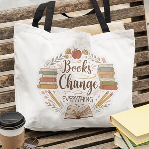 May include: White tote bag with black handles featuring the text "Books Change Everything" surrounded by illustrations of books, pencils, and an apple. A coffee cup, sunglasses, and books are next to the bag.