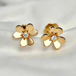 May include: A pair of gold stud earrings shaped like four-leaf clovers. Each earring features a polished gold finish with a single, small, clear, round-cut gemstone set in the center of the flower petals, creating a bright and reflective appearance.