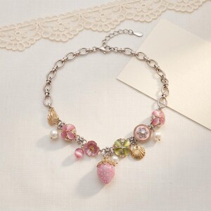 May include: A silver-toned necklace featuring pink floral and strawberry charms, along with faux pearls and a seashell pendant. The necklace has a delicate chain and a lobster clasp closure. The design includes a variety of pink, green, and gold accents.