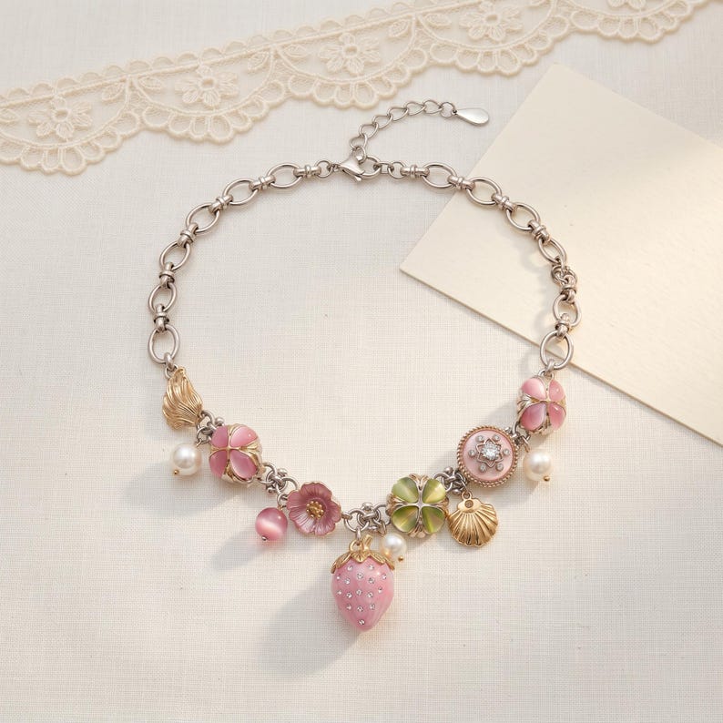 May include: A silver-toned necklace featuring pink floral and strawberry charms, along with faux pearls and a seashell pendant. The necklace has a delicate chain and a lobster clasp closure. The design includes a variety of pink, green, and gold accents.