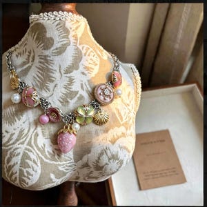 May include: A silver-toned necklace with pink and gold accents, featuring strawberry, flower, and seashell charms. The necklace includes faux pearls and a decorative pendant with a central gemstone. The necklace is displayed on a mannequin.